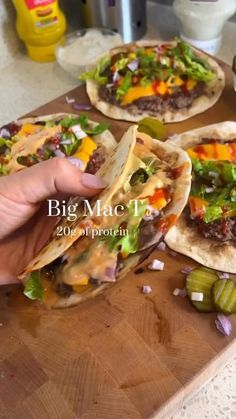 three tacos on a cutting board with pickles and other food items in the background