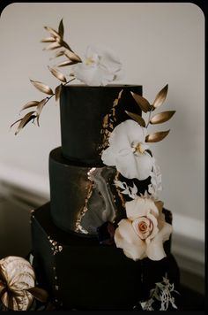 a three tiered black cake with white flowers and gold leaf decorations on it's side