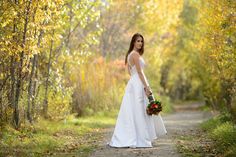 a woman in a white wedding dress is standing on a dirt road surrounded by trees