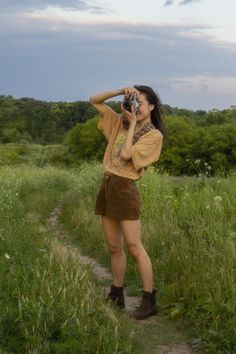 A girl standing in a field taking a photo with a film camera. She is wearing a tan graphic tee, brown corduroy shorts, and brown boots. Loose Earthy Outfits, Summer Outfits Earth Tones, Earth Tones Summer Outfits, Earth Tones Wardrobe, Earthy Going Out Outfit, Granola Lesbian Style, Casual Earth Tone Outfits, Earthy Tone Outfits Aesthetic, Granola Fashion Aesthetic