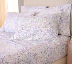 a bed with white sheets and purple flowers on the comforter next to a night stand