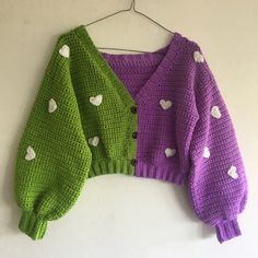 two knitted sweaters hanging up on a white wall, one has hearts and the other is green