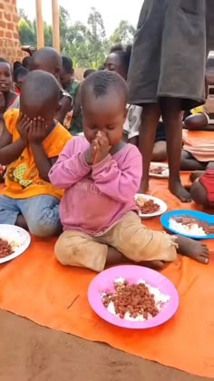 several children sitting on the ground with plates of food in front of them and one child eating