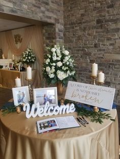 a welcome table with flowers, candles and pictures on it in front of a brick wall