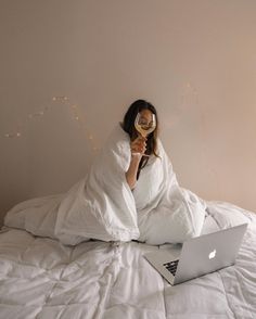 a woman laying in bed with a laptop and wine glass on her face as she looks at the screen