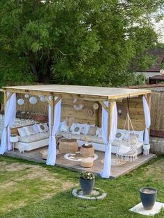 a wooden gazebo with white curtains and pillows on it in the grass next to trees