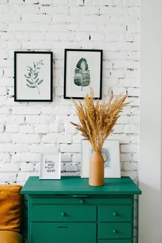 a green dresser with two pictures on the wall above it and a plant in a vase