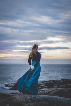 a woman in a blue dress standing on rocks near the ocean with her back to the camera