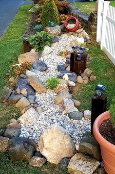 a garden with rocks and plants next to a white picket fence on the grass near a house