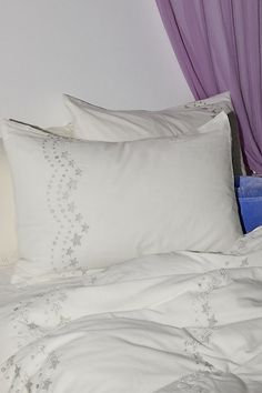 a white bed topped with lots of pillows next to a purple drapeed window