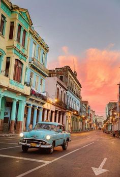 an old car is driving down the street in front of some colorful buildings at sunset