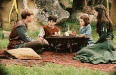 four children sitting on a blanket talking to each other in front of a picnic table