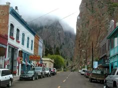 cars parked on the side of a road in front of buildings and mountains with clouds