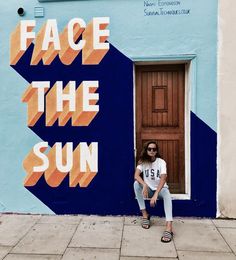 a woman sitting on the ground in front of a blue wall with an orange and white sign that says face the sun