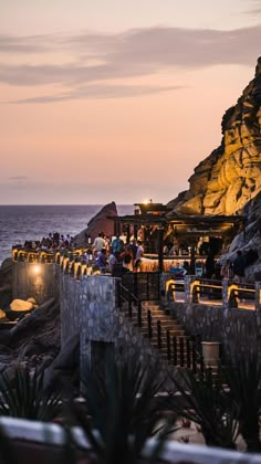 people are sitting at tables on the beach by the water and cliffs in the background