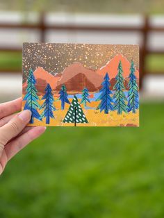 a hand holding up a card with trees on it in front of a fence and mountains