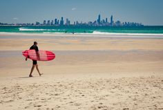 a man with a surfboard walks on the beach in front of the city skyline