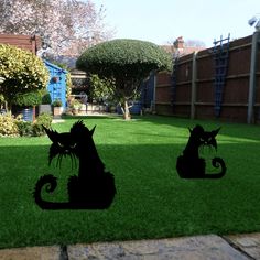 two silhouettes of cats sitting on the grass in front of a fenced back yard