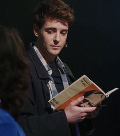 a young man reading a book while standing in front of a woman with dark hair