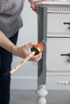 a woman is painting an old dresser with white paint and a wooden brush in her hand