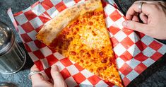 a slice of cheese pizza on a red and white checkered paper with two hands