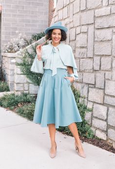 a woman standing in front of a brick wall wearing a blue dress and matching hat