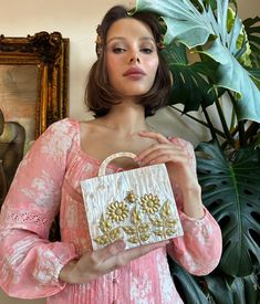 a woman holding a purse in front of a painting and potted plant behind her