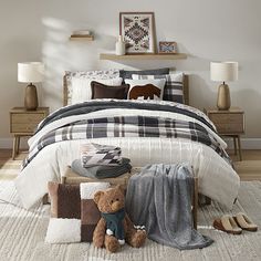 a teddy bear sitting on the floor in front of a bed with pillows and blankets