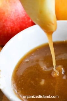 someone pouring caramel sauce into a white bowl with apples and oranges in the background