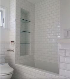 a white toilet sitting next to a bath tub under a window in a room with white tiles on the walls