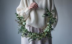 a woman is holding a wreath with greenery on it while wearing a white sweater