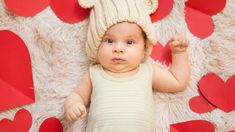 a baby wearing a knitted bear hat laying on a rug with red paper hearts