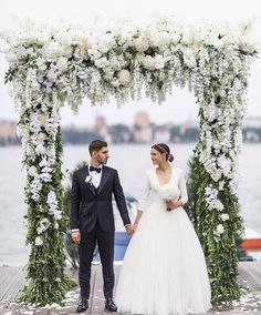 a bride and groom holding hands under an arch with flowers on it near the water