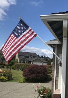 an american flag flying in the wind on a house's front yard with flowers and shrubs