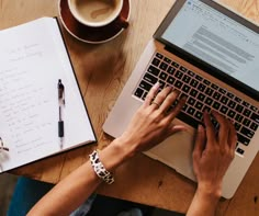 a person typing on a laptop computer next to a cup of coffee and notebooks