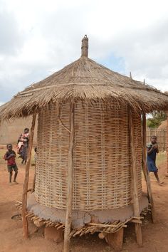 a hut made out of straw with people standing around it on dirt ground near by