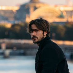 a man with long hair standing in front of a body of water and looking off into the distance