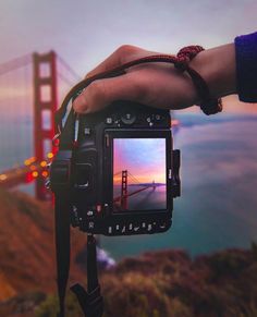 a person holding up a camera with the golden gate bridge in the background at sunset