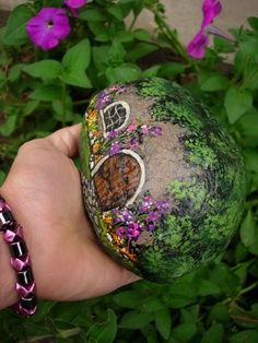 a hand holding a rock with a house on it and flowers in the back ground