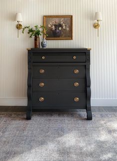 a black dresser with gold handles and knobs in a white walled room next to a painting on the wall