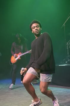 a man standing on stage with a guitar in his hand and another person holding a microphone behind him