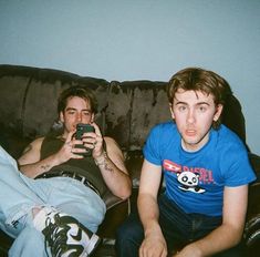 two young men sitting on a couch looking at their cell phones while the man is taking a selfie