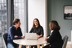 three people are sitting at a table with a laptop in front of them, talking