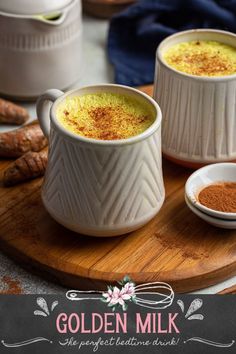 two white mugs filled with golden milk sitting on top of a wooden cutting board