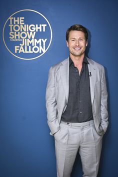 a man in a gray suit and black shirt standing next to a blue wall with the tonight show jimmy falcon logo on it