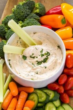 a bowl of dip surrounded by veggies on a cutting board with carrots, celery and peppers