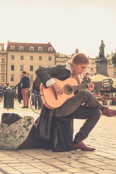 a man sitting on the ground playing an acoustic guitar in front of a building with people walking around