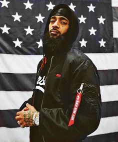 a man standing in front of an american flag wearing a black jacket and hoodie