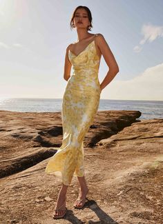 a woman in a yellow dress standing on the rocks by the ocean with her hands on her hips