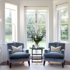 two chairs and a table in front of three windows with blue couches, one is facing the other